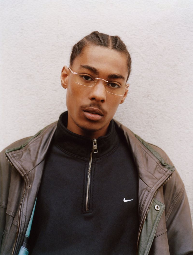 Close-up portrait of a Black man with braided hair wearing rimless glasses, a black zip-up top, and a brown leather jacket.