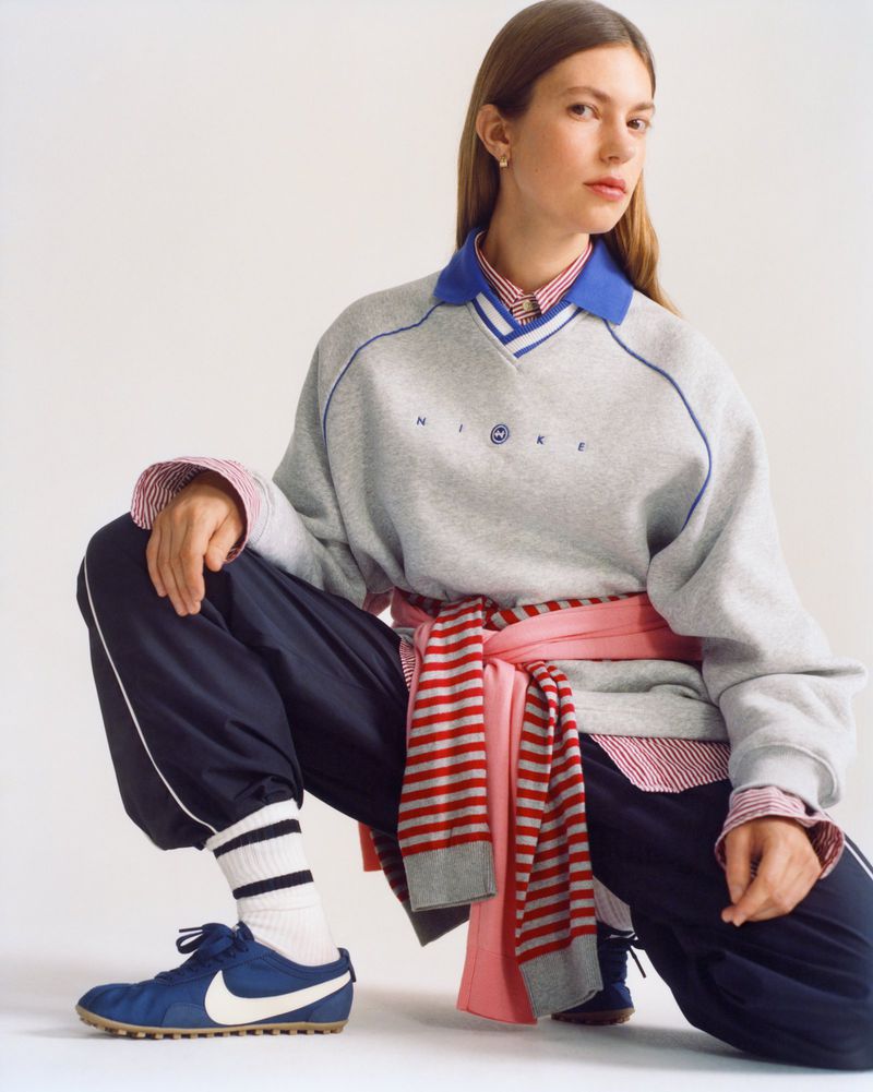 Model in grey sweatshirt and navy track pants squatting against a white studio background for Nike SS26.