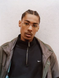 Close-up portrait of a Black man with braided hair wearing rimless glasses, a black zip-up top, and a brown leather jacket.