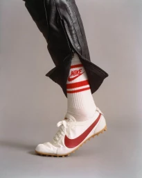 Close-up shot of a white sneaker with red swoosh, striped Nike socks, and black leather trousers against a grey seamless background