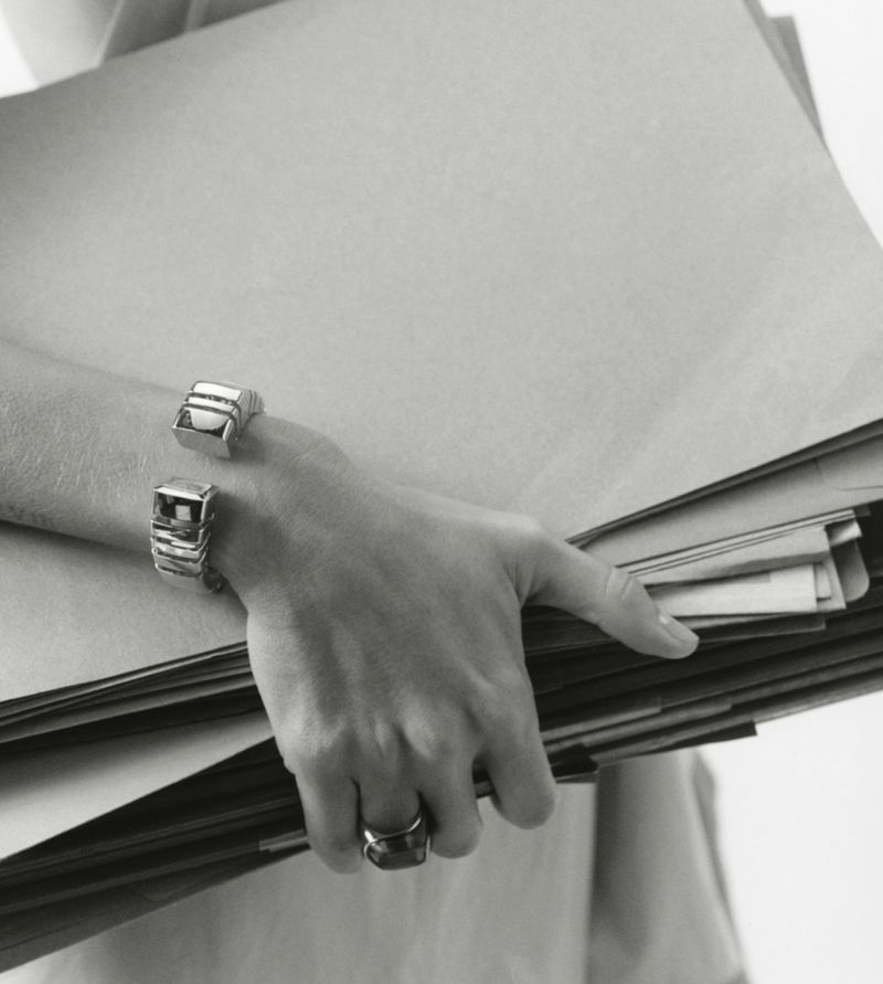 Close-up shot of a person holding a stack of brown papers with silver ring and bracelet accessories in a monochrome light.