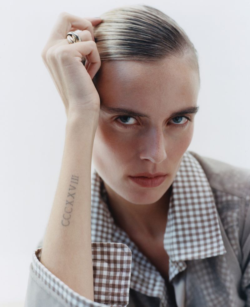 Close-up studio portrait of a serious adult female model with blonde hair, a ring, visible tattoo, and a checkered collared shirt.