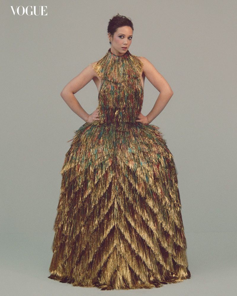 Fashion photo of an adult woman in a golden dress posing in front of a plain background, showcasing haute couture style and Vogue magazine.