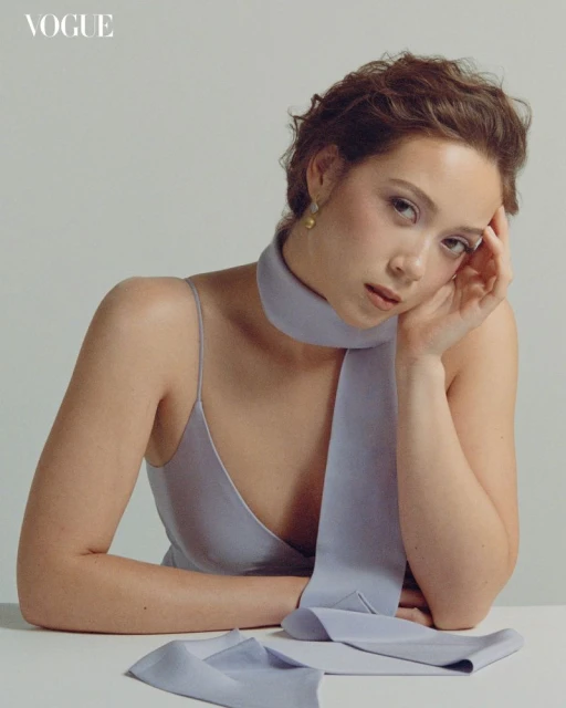 Close-up studio portrait of a young woman with short curly hair, wearing a light purple dress and scarf, resting her head on her hand.