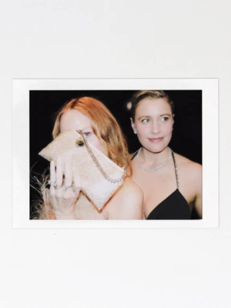 Two women posing together, one holding a sparkling clutch bag to partially hide her face while the other smiles gently beside her.