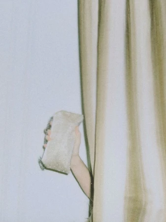 Hand holding a textured light-colored handbag while peeking out from behind draped curtains against a plain wall