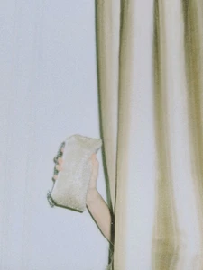 Hand holding a textured light-colored handbag while peeking out from behind draped curtains against a plain wall