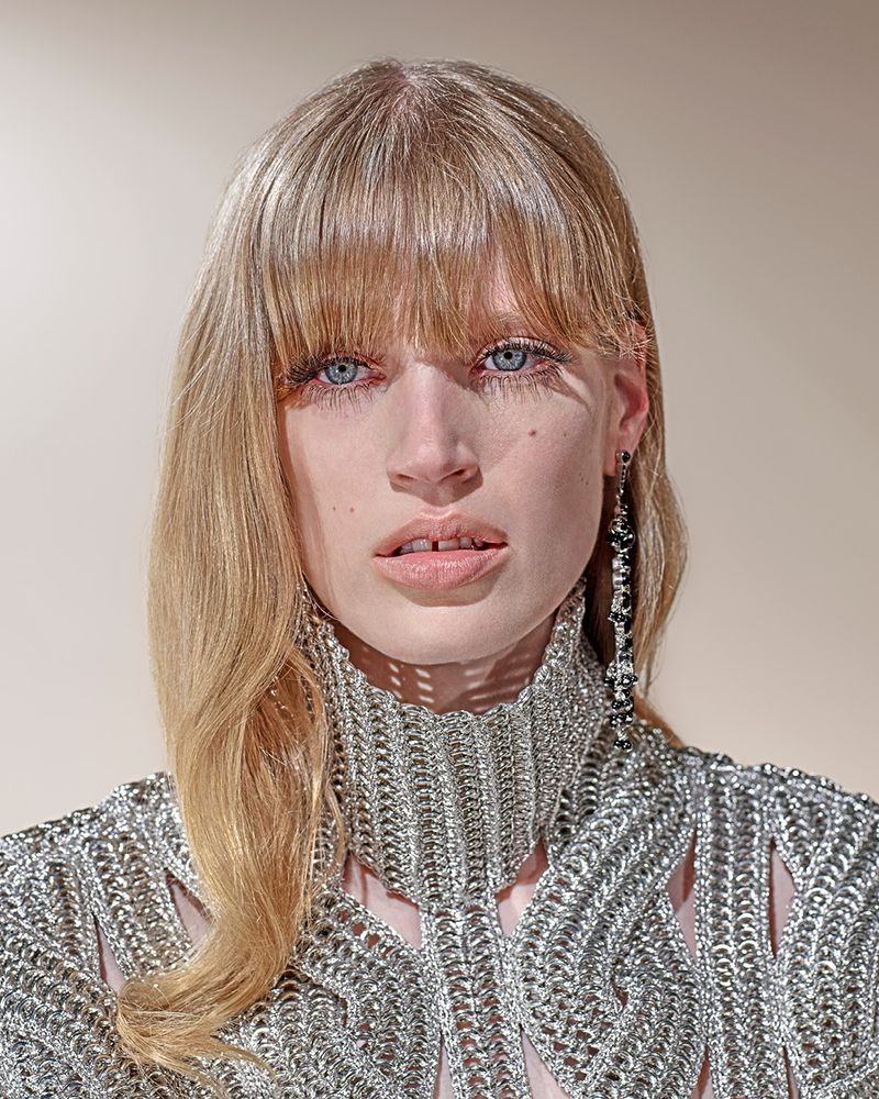 Close-up portrait of a blonde woman wearing a silver metallic high-necked garment and hanging earrings against a light beige background.