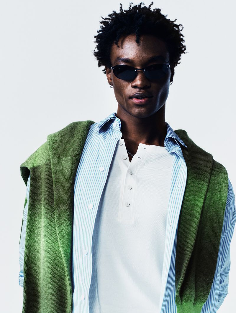 Stylish African American man in sunglasses, striped shirt, and green knit sweater, posing in a bright photo studio. Fashion shot.