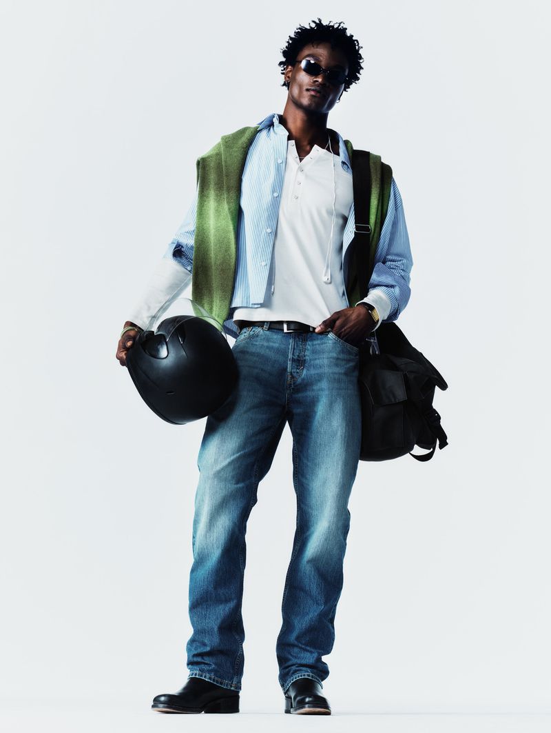 Black man holding a black helmet and wearing a white polo shirt, blue jeans, and a green sweater draped over his shoulders.