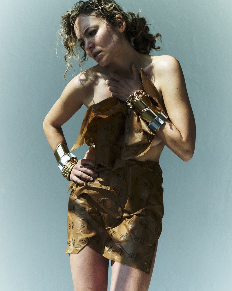 Woman wearing brown wrapping paper garment and chunky gold bracelets against a plain blue background.