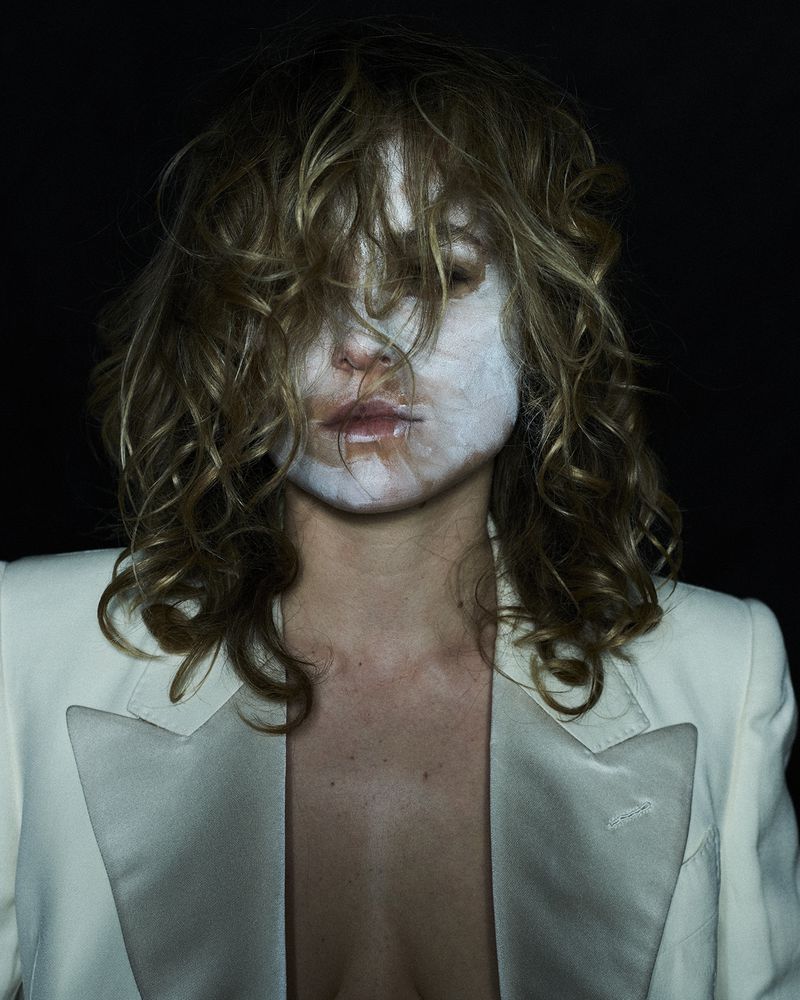 Portrait of a woman with white face makeup wearing a white blazer against a dark background