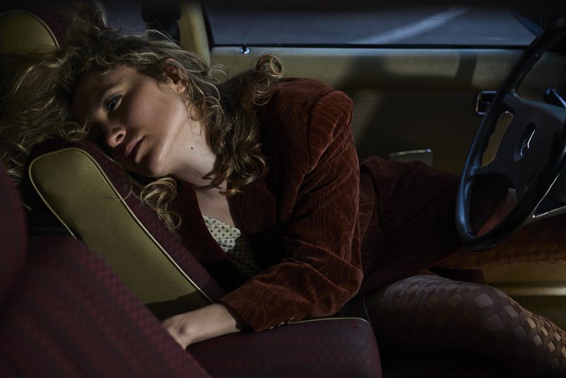 Woman with blonde curly hair wearing a brown velvet blazer lying across the front seat of a car