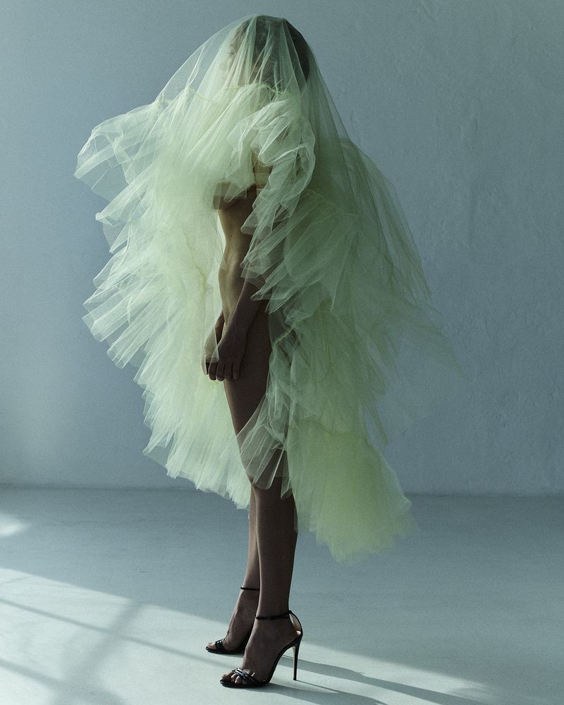 Model standing in profile wearing a voluminous pale green tulle garment and black high-heeled sandals in a minimalist interior.