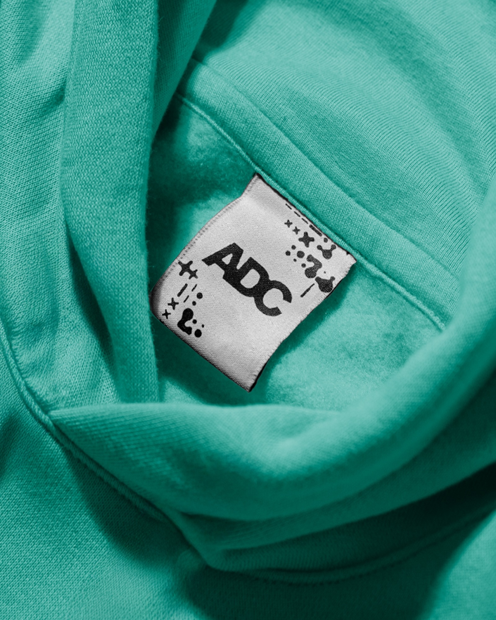 Close-up of a teal hoodie collar showing a white branded clothing tag with ADC printed in bold black letters
