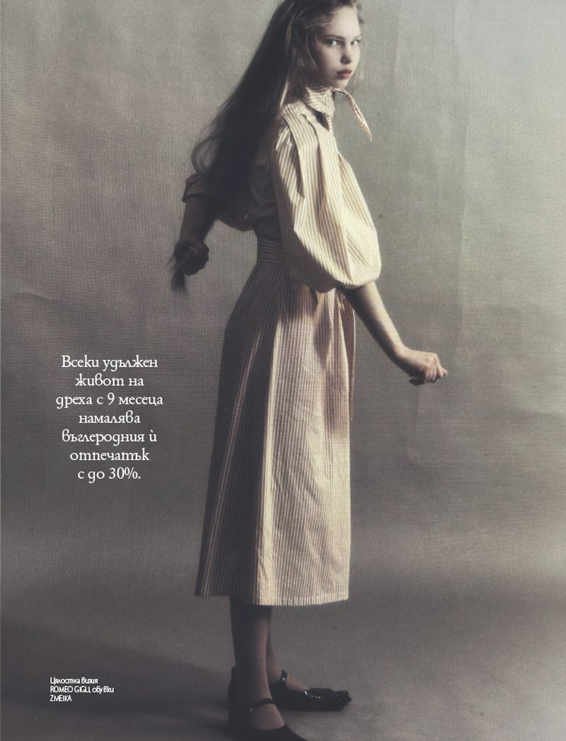 Young woman in a striped dress strikes a pose in a fashion editorial shot against a textured backdrop, with long hair and minimal makeup.