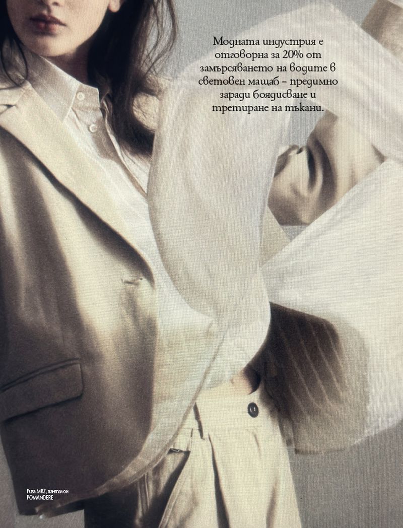 A woman poses in a tan pantsuit with a collared shirt. The photo is a close crop of the subject in muted light.