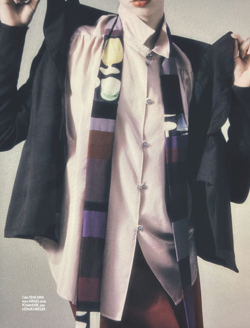Close-up studio fashion shot of a slender adult wearing a pale pink button-down shirt, patterned scarf, and black jacket.