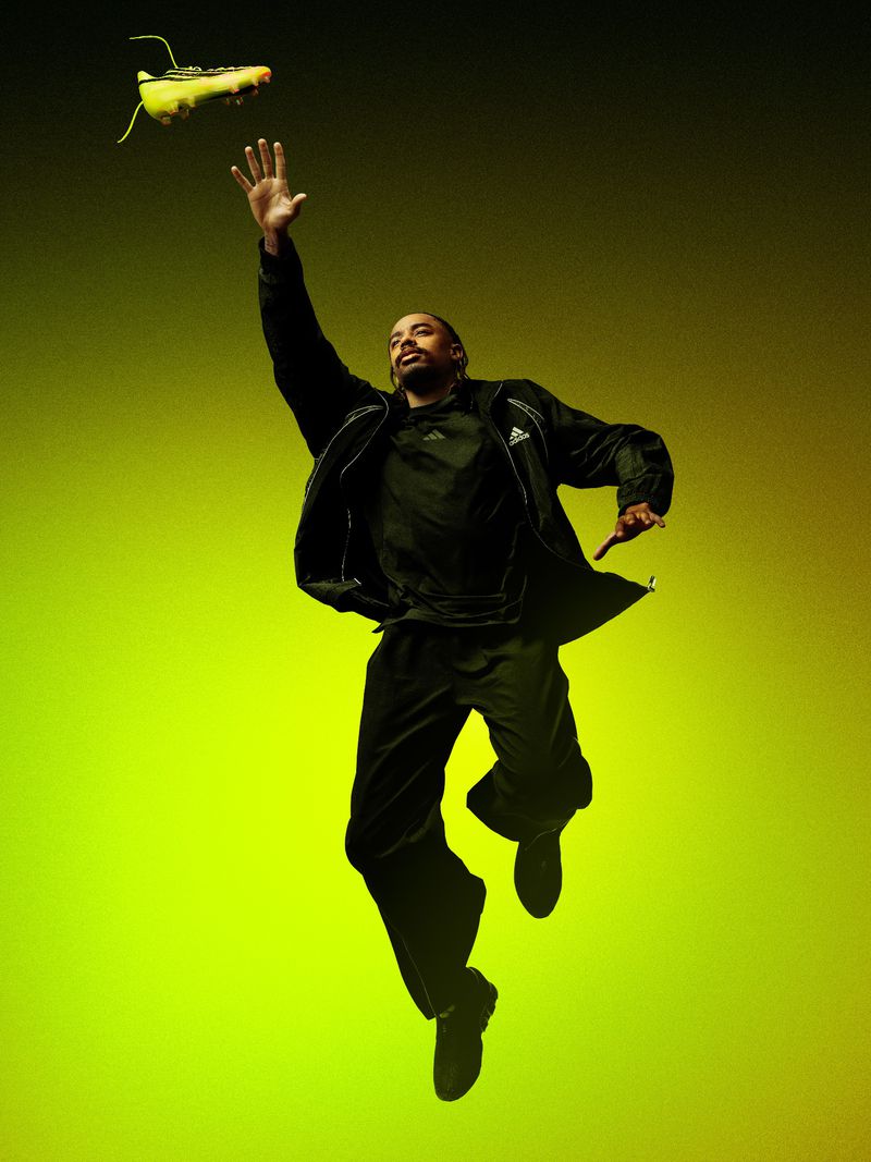 Man leaping in the air reaching for a neon yellow football boot against a green and yellow gradient studio background.