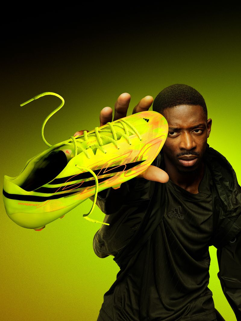 Close-up portrait of a Black man holding a bright yellow and orange Adidas F50 football boot toward the camera against a green background