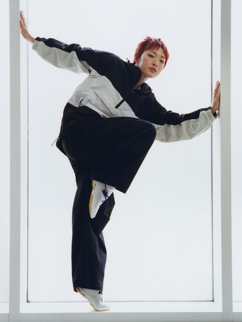An adult female with red hair poses indoors near a window wearing a color-blocked jacket and dark pants in a fashion studio shot.