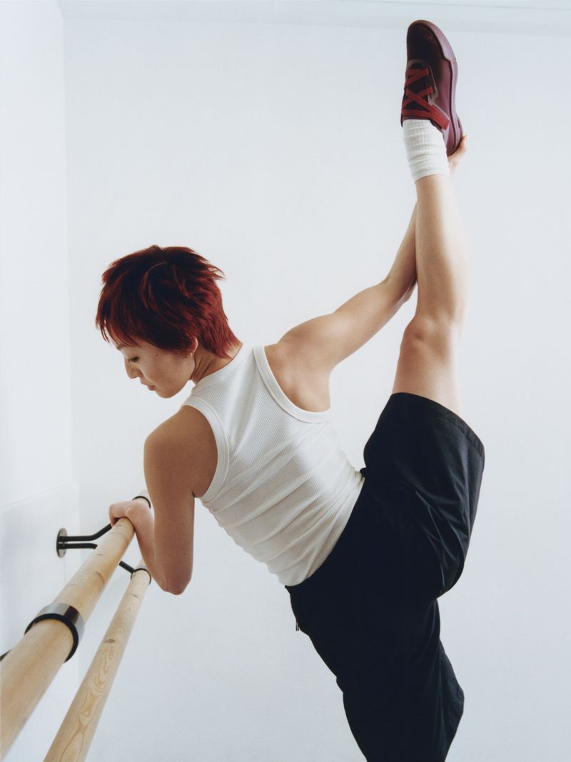 Woman in white tank top and black shorts holding her leg high while leaning on a wooden ballet barre against a white wall.