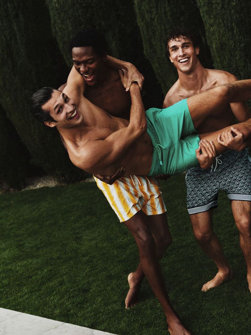 Three male models laughing outdoors, with one being held up playfully against a dark cypress hedge backdrop
