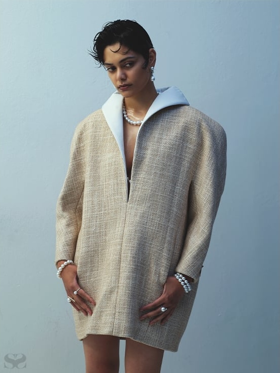 Woman posing in a tan, oversized coat, pearl jewelry, with a soft blue background in a studio fashion photograph. Minimalist aesthetic.