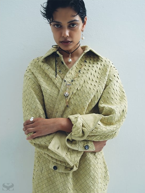 An adult woman wearing a light yellow woven-texture collared shirt and jewelry poses against a white wall in a fashion shot.