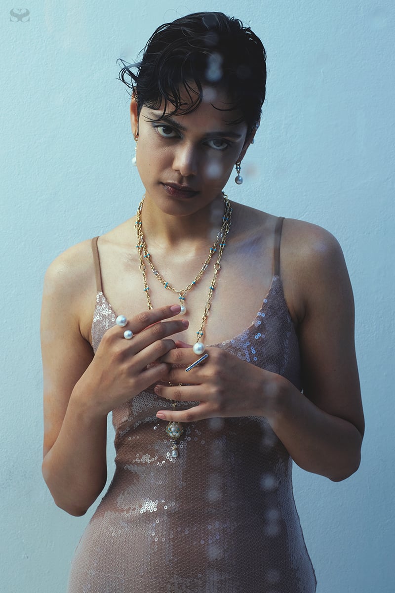 A young adult woman with short dark hair and neutral expression poses indoors wearing a sequined dress and pearl jewelry in a portrait shot.