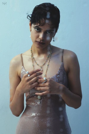 A young adult woman with short dark hair and neutral expression poses indoors wearing a sequined dress and pearl jewelry in a portrait shot.