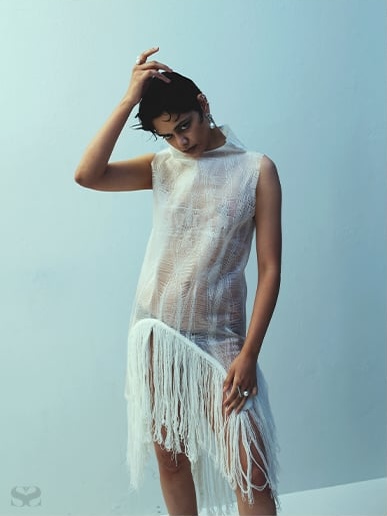 Model with short dark hair wearing a sheer white sleeveless dress with fringe detail against a pale blue studio background