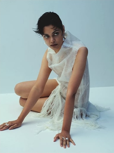 A woman with dark hair sits in a white fringe dress and pearl jewelry, gazing upward in a studio setting against a pale backdrop.