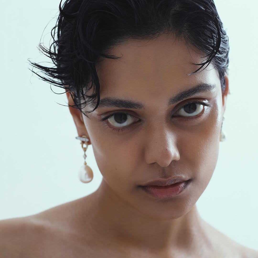 Close-up portrait of a young woman with short, slicked-back dark hair, wearing pearl drop earrings against a light, neutral backdrop.
