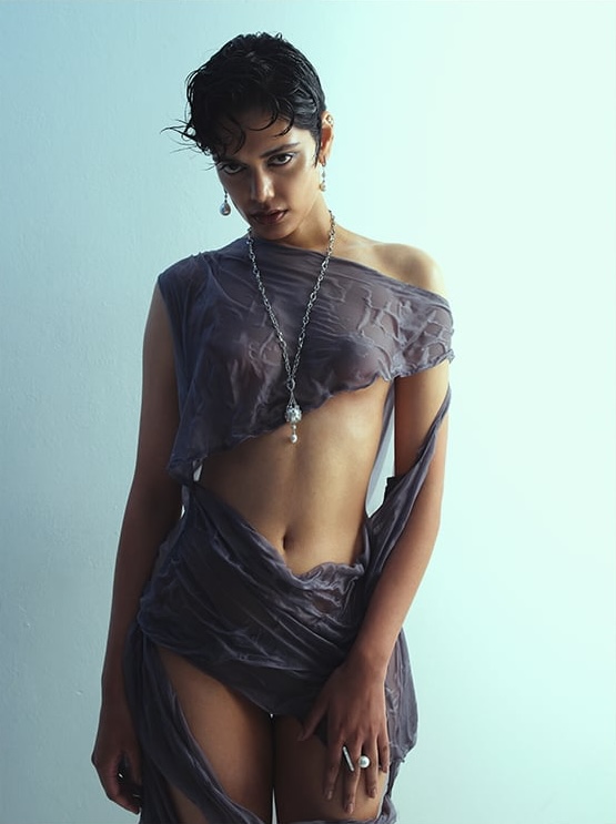 Young woman poses in draped grey fabric with jewelry on light blue background in studio setting, fashion, beauty, and portrait photography.