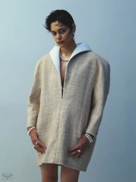 Model with short dark hair wearing a textured beige coat with white collar and pearl jewelry against a plain grey wall.