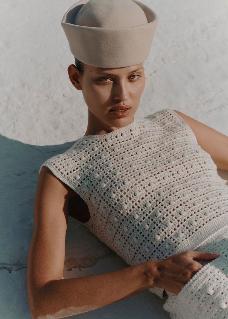 Woman in a knitted top and sailor hat poses against a neutral wall in sunlit, medium shot with soft shadows and natural skin tones.