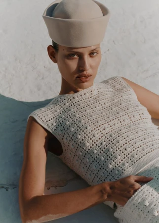 Woman in a knitted top and sailor hat poses against a neutral wall in sunlit, medium shot with soft shadows and natural skin tones.