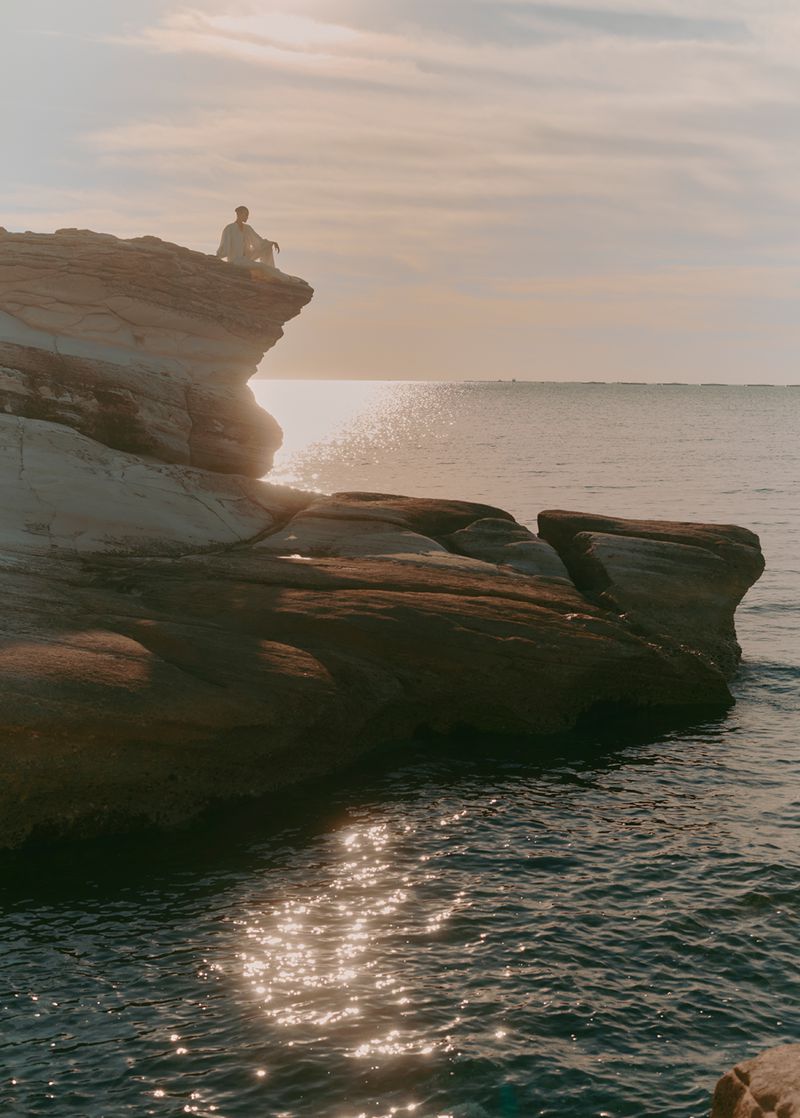 Person sitting on rocky cliff overlooking ocean water at sunset. Calm outdoor scene with peaceful mood.