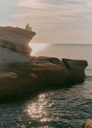 Person sitting on rocky cliff overlooking ocean water at sunset. Calm outdoor scene with peaceful mood.