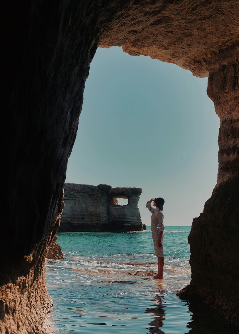 Person in white stands in ocean water, framed by a rock cave, facing a rock formation with a hole, under a bright blue sky.