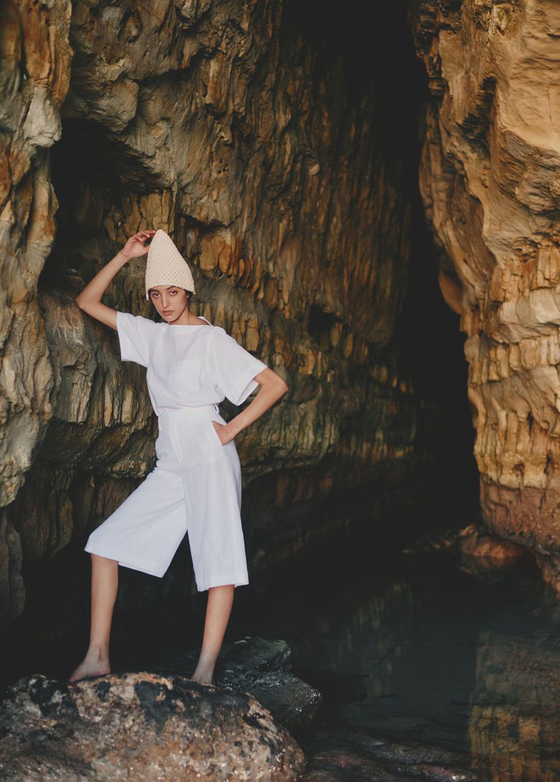 A woman in white culottes and a short-sleeve shirt stands in a rock cave, wearing a knit hat, against a background of brown and beige stone.