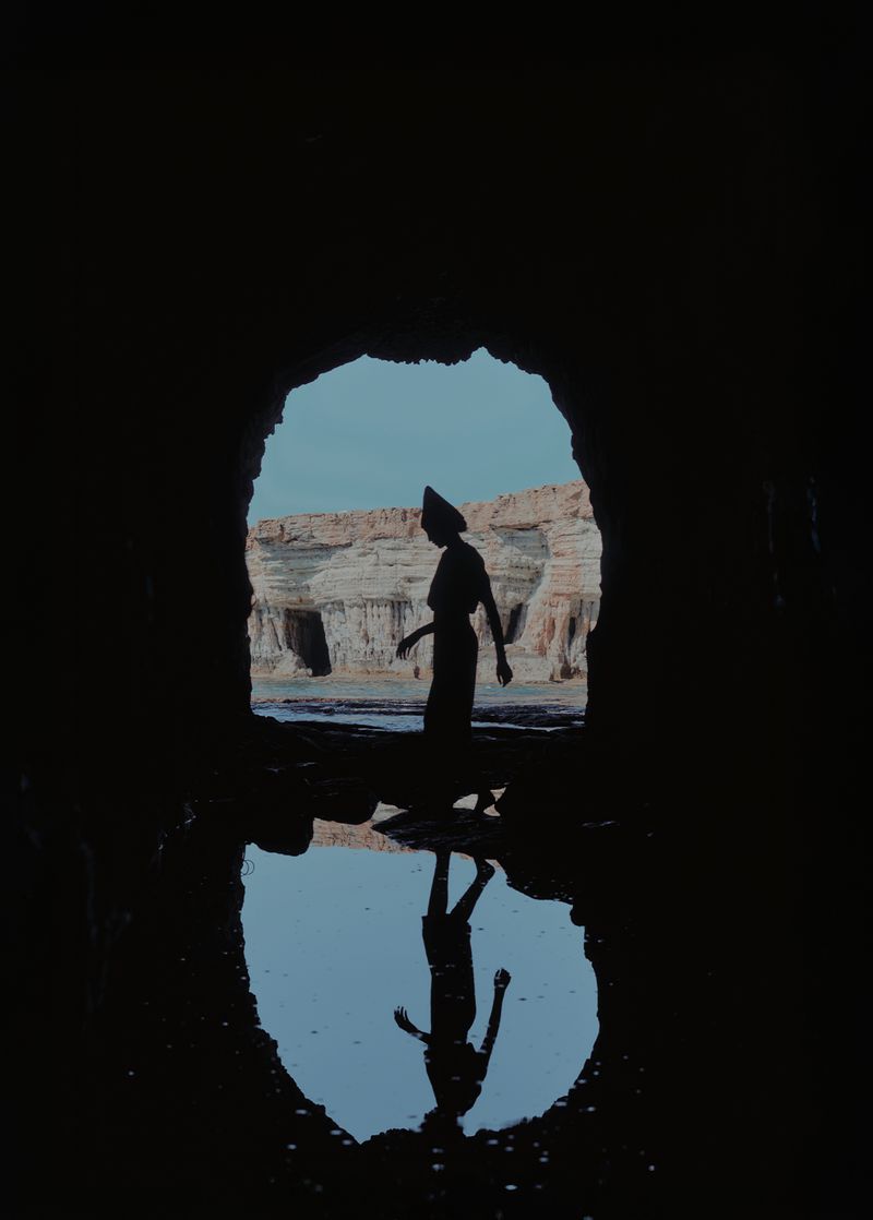 Silhouette of a woman walking through a cave opening overlooking rock formations and water, reflection visible in foreground.