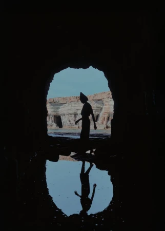 Silhouette of a woman walking through a cave opening overlooking rock formations and water, reflection visible in foreground.