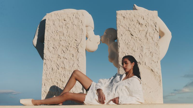 An adult female model poses in a white outfit, lying on a stone surface, with a massive kissing sculpture behind her under the sunlight.