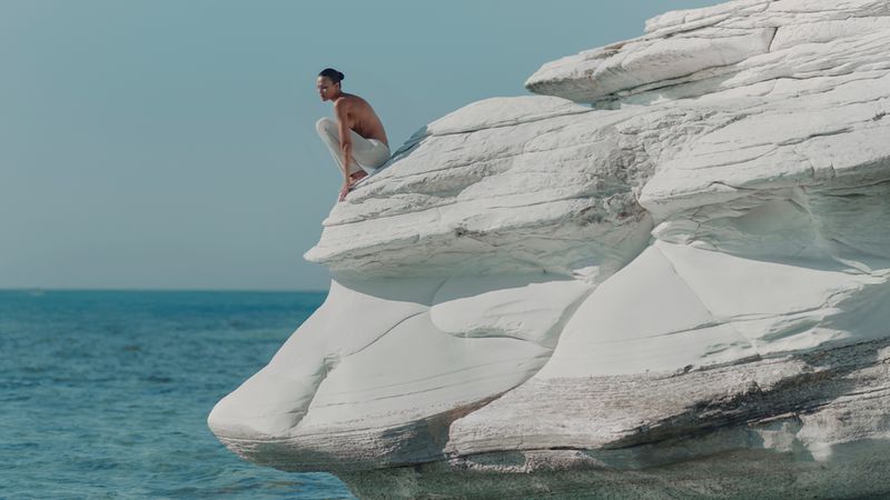 Man sitting on white rock formation near ocean. The man is wearing white pants, with a blue sky and ocean in the background.