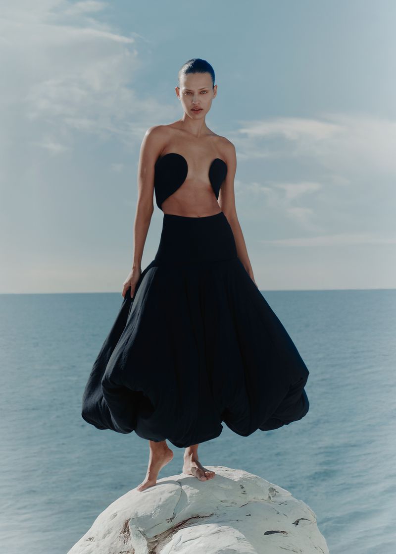 Model in a black strapless cutout dress with a voluminous balloon skirt standing barefoot on a white rock above the sea.