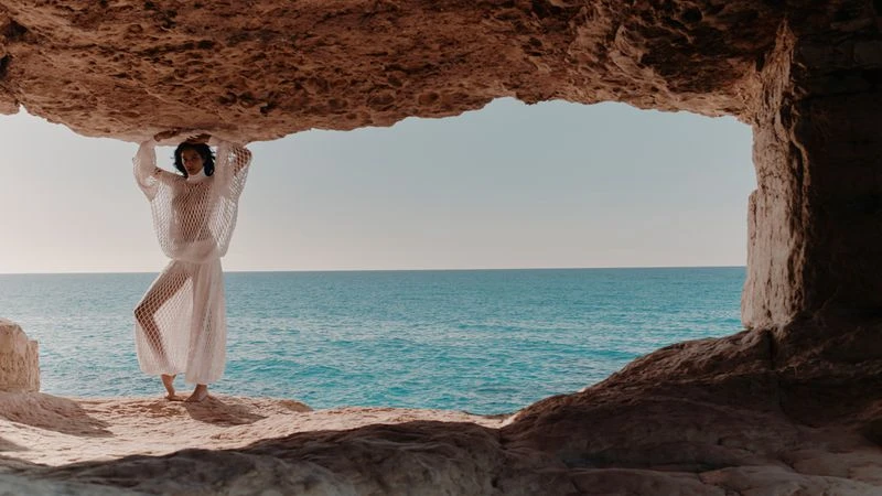Model posing in a sheer white net outfit inside a natural rock cave framing the calm turquoise ocean horizon.