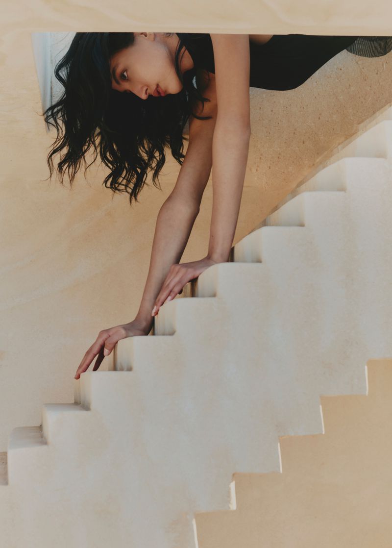 Woman with long dark hair in a black top reaches down a white plaster staircase in an architectural portrait with soft lighting.