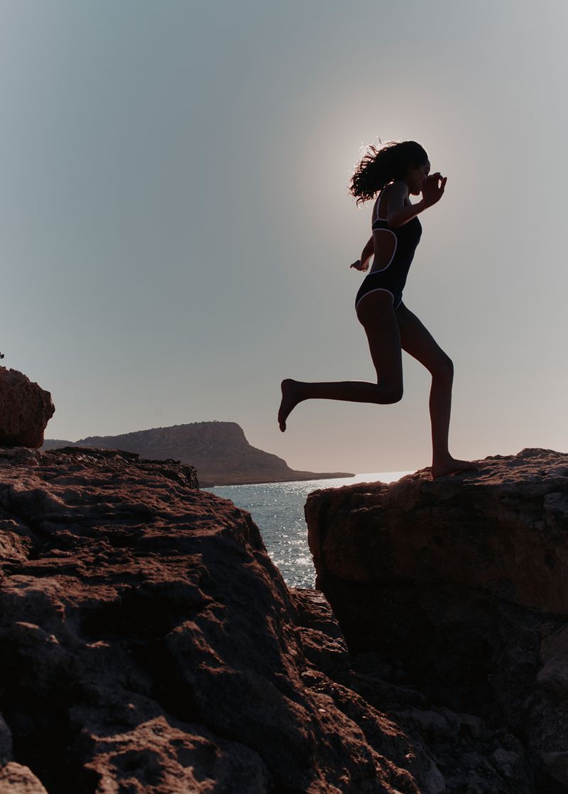 Silhouette of a woman leaping across rocky coastal cliffs against the bright backlit sun over the sea.
